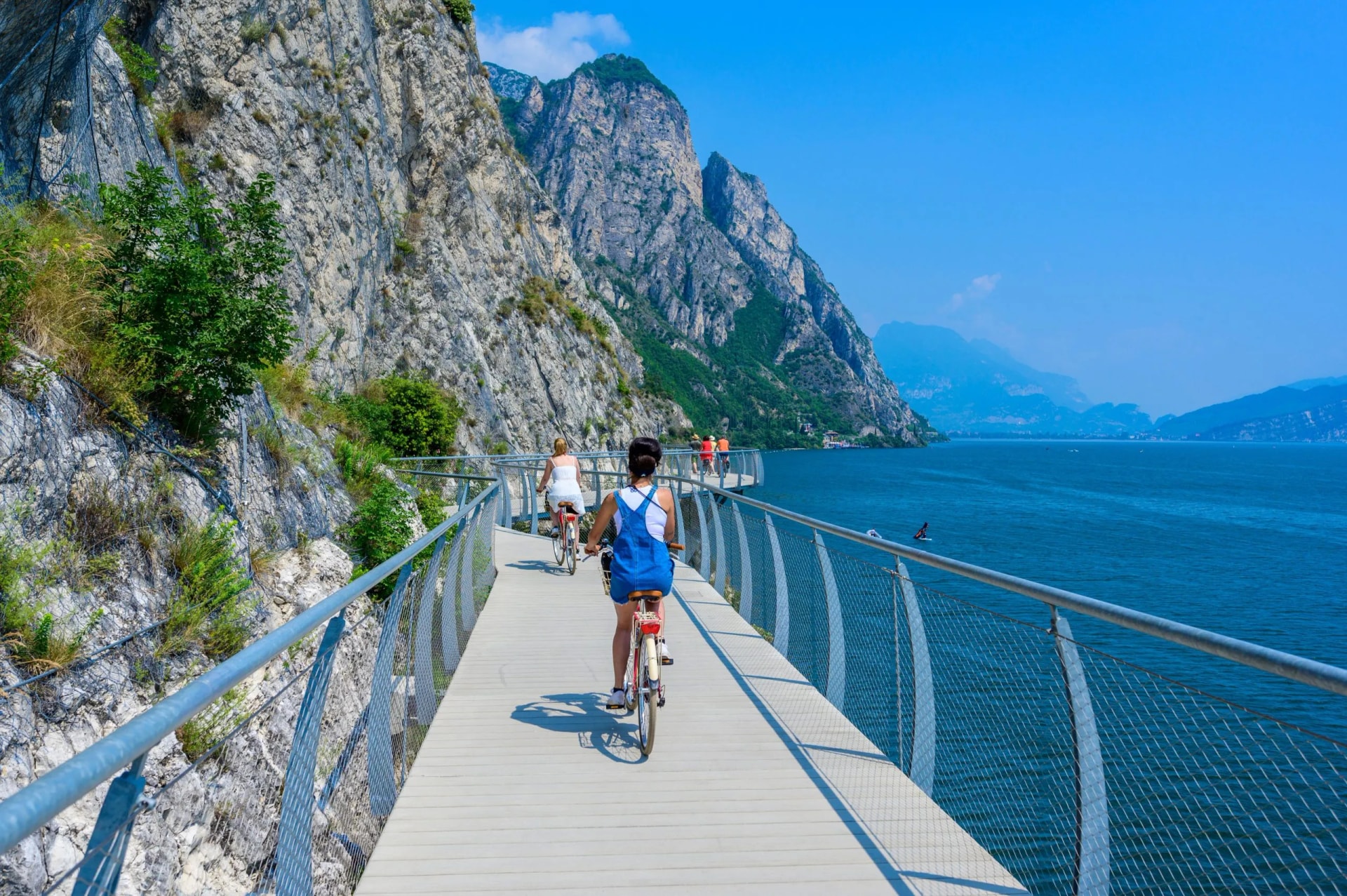 Cyclists on a suspended bikeway alongside a steep cliff overlooking bright blue water, Garda.