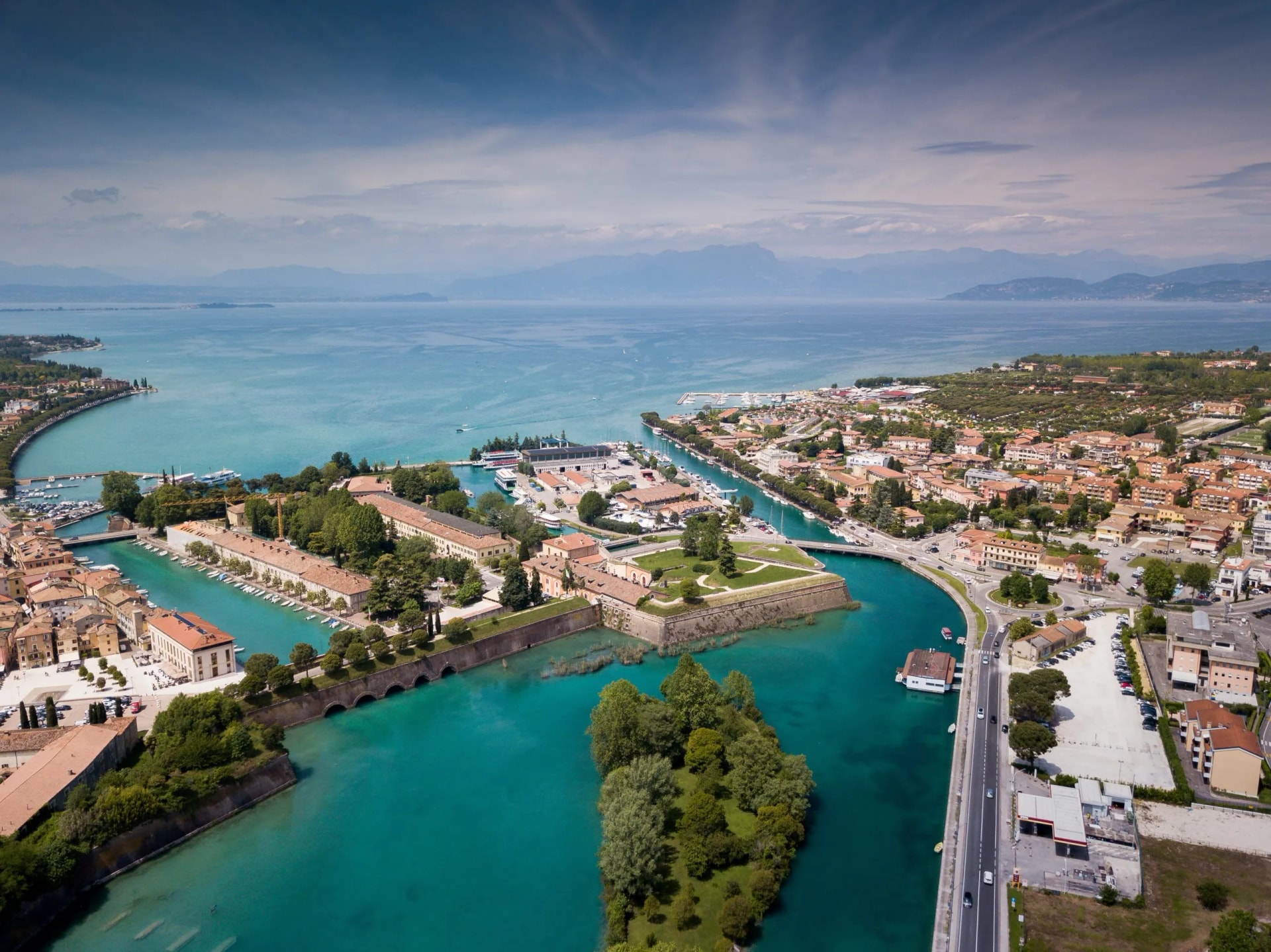 Aerial view of Peschiera town, harbor, and turquoise water meeting a large lake with mountains in the distance.