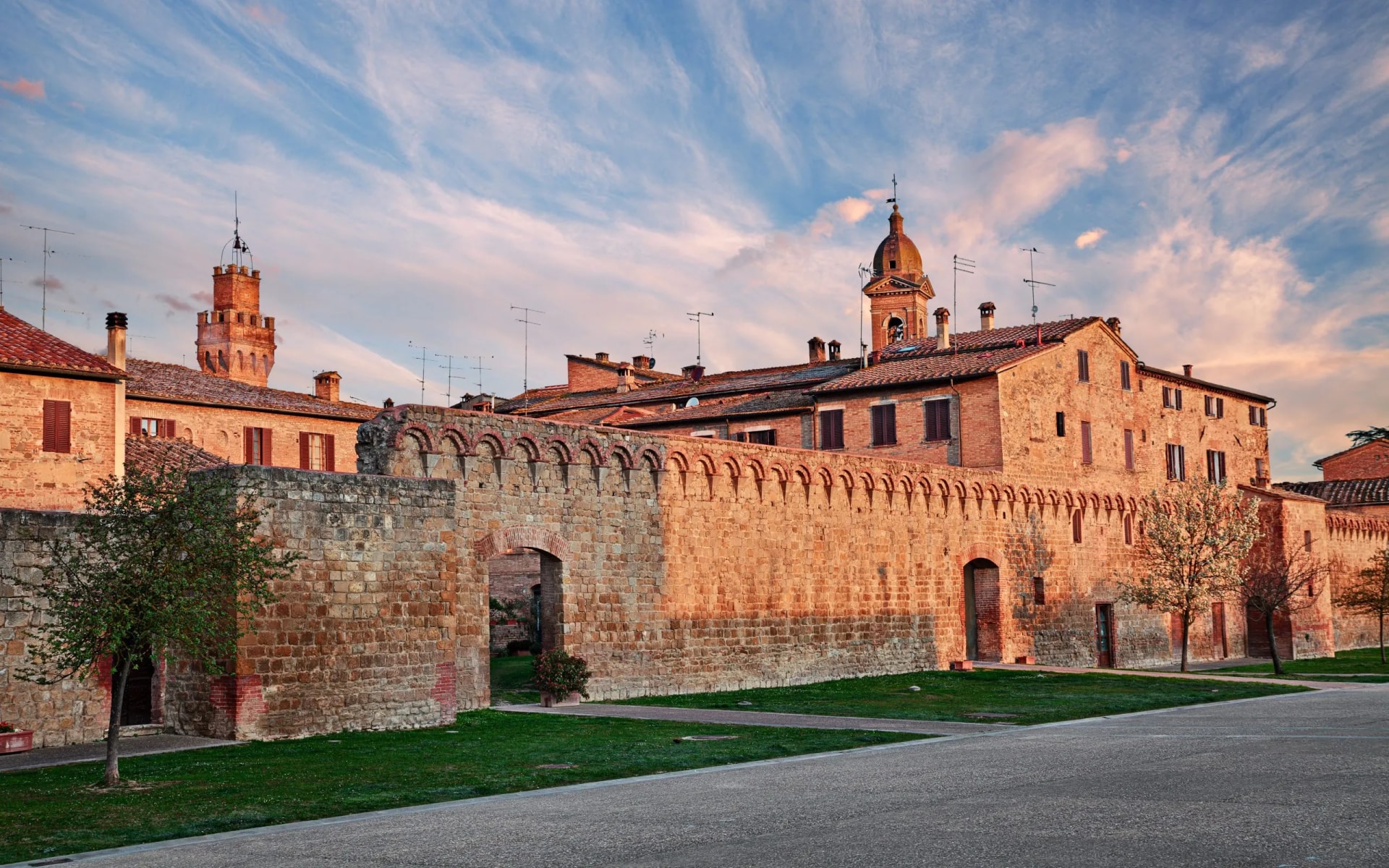 Historic stone wall and brick buildings illuminated by sunset in Buonconvento under a blue, cloudy sky.
