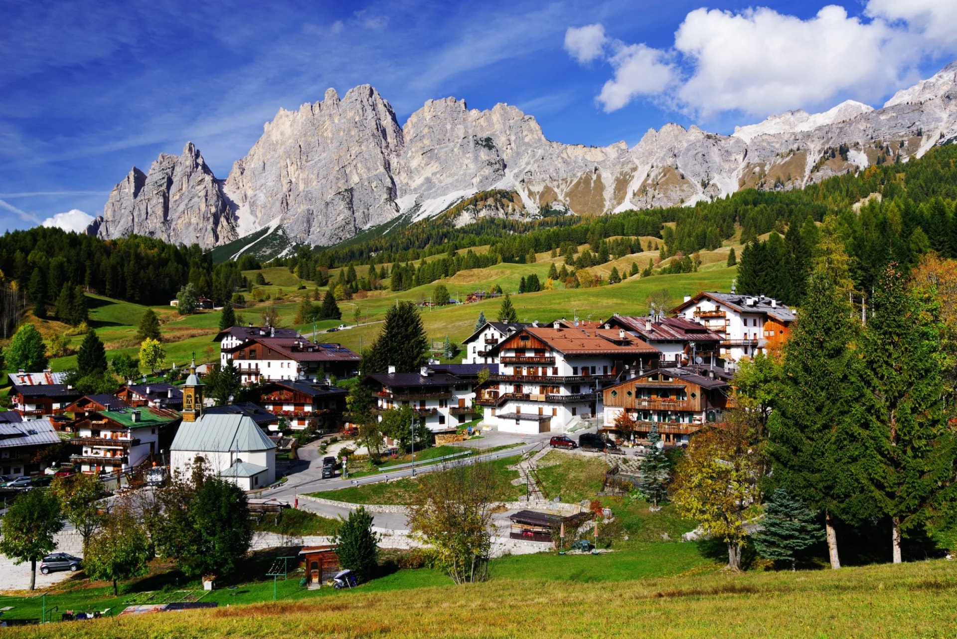 Alpine village with traditional chalets nestled below jagged, rocky mountains under a blue sky.