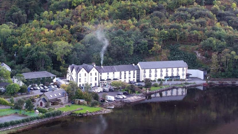 White hotel building by dark water with smoke rising, backed by dense green hillside.