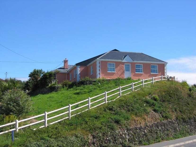 Pink cottage on grassy hill with white fence under bright blue sky, Cottage Heights B&B