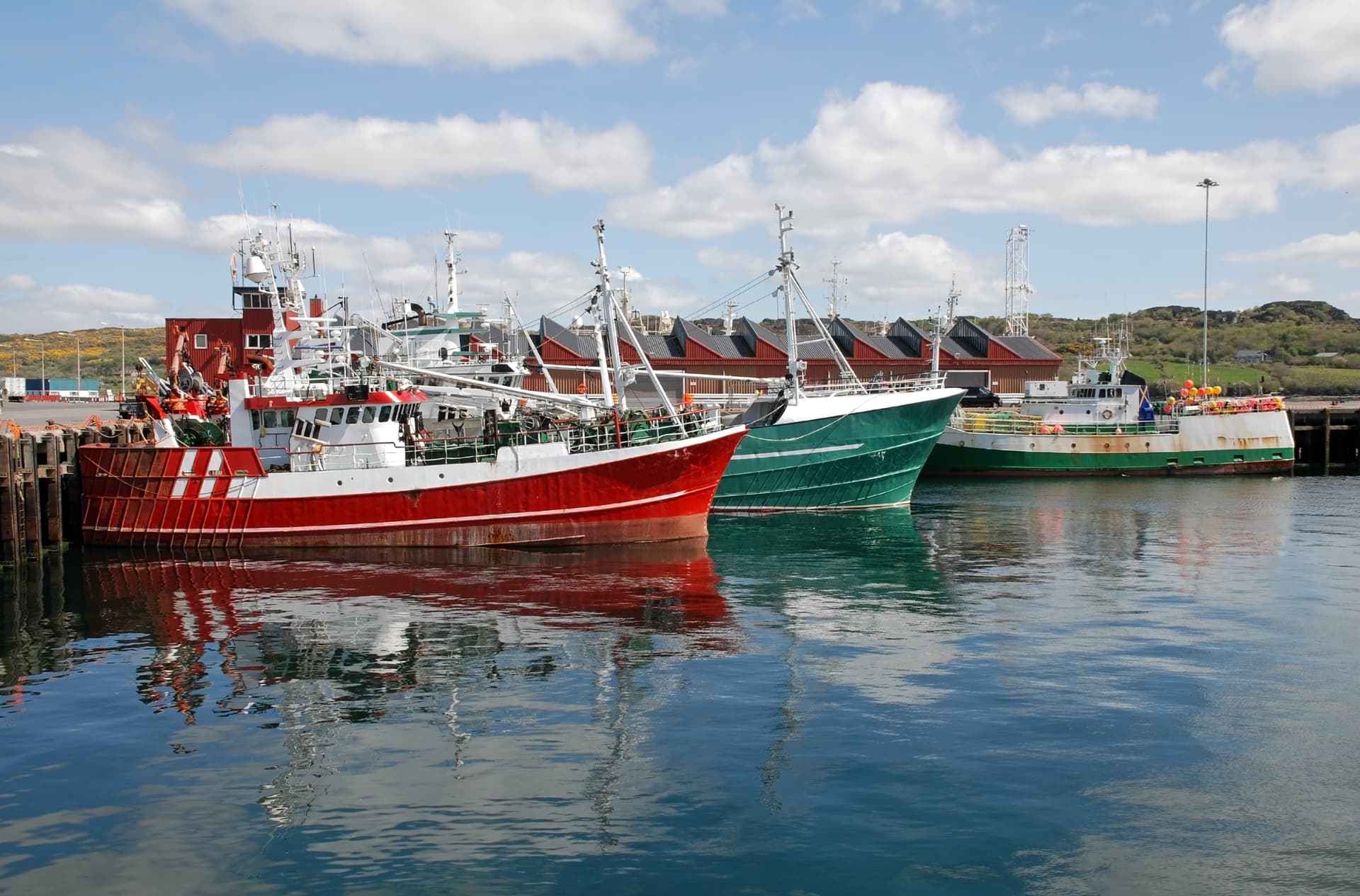 Fishing ships docked in Killybegs harbor with reflections on the blue water under a cloudy sky.