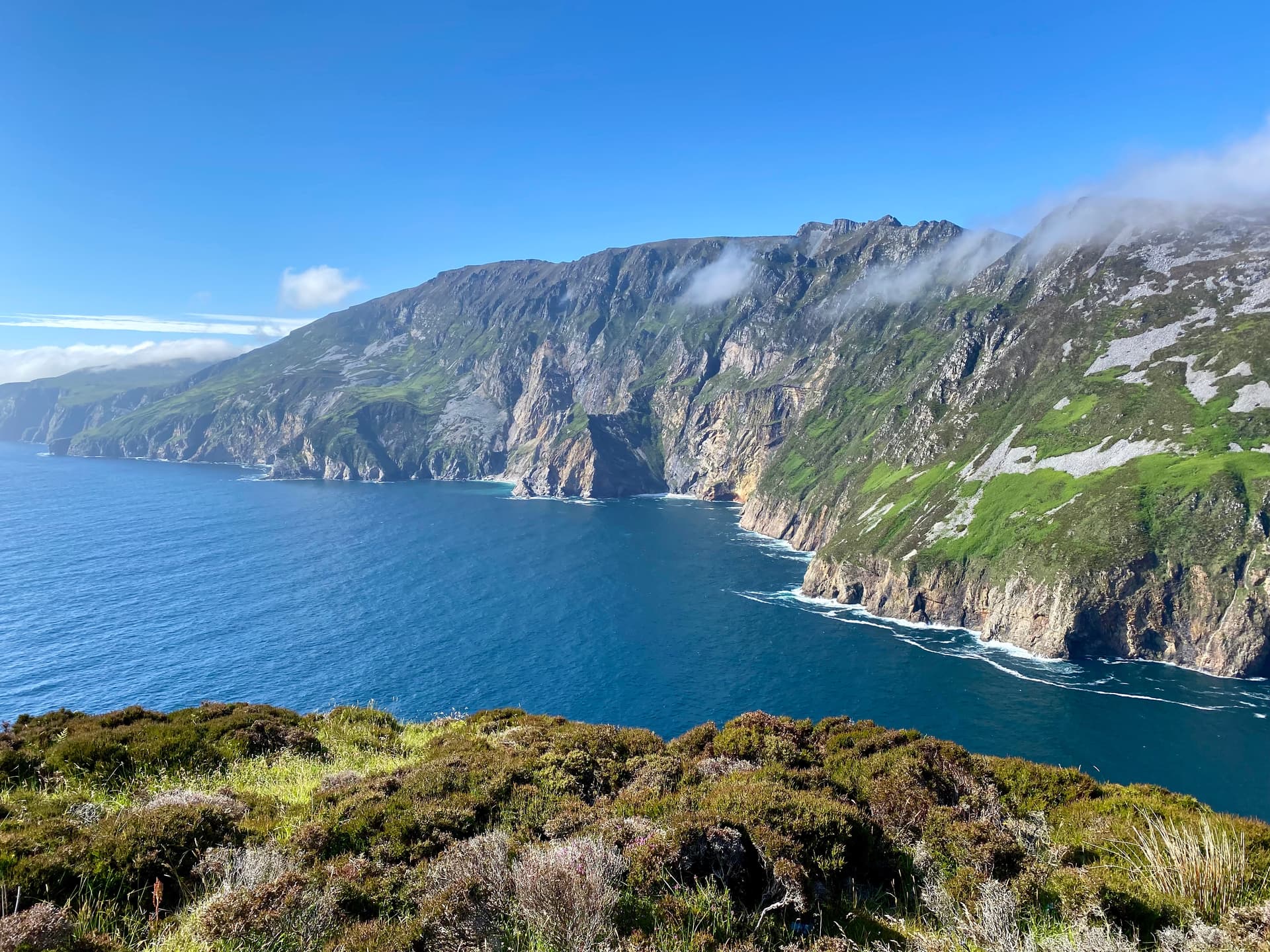 Sliabh Liag sea cliffs meeting deep blue water under a bright blue sky with low clouds.