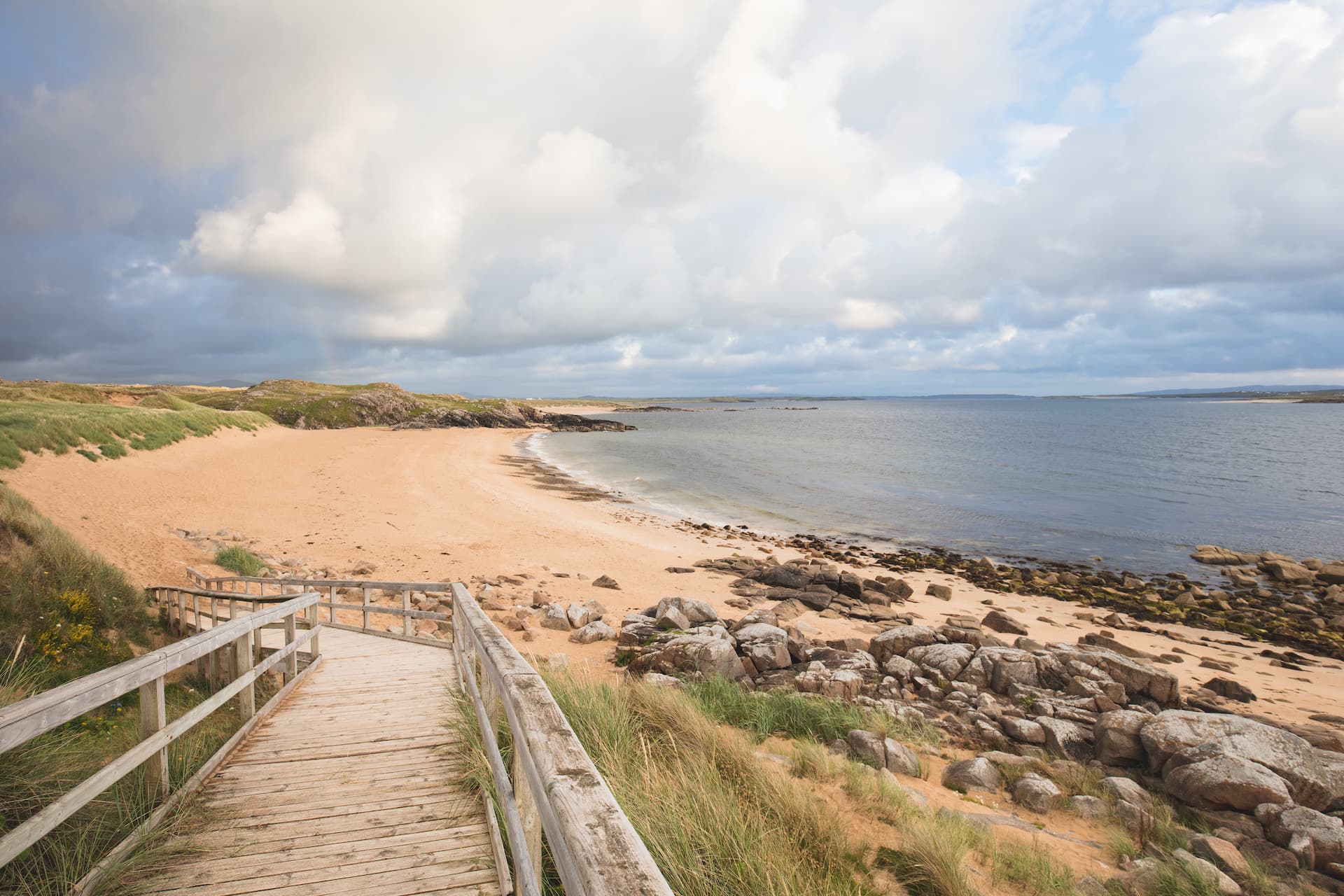Wooden boardwalk leading to Maghera Beach with sand dunes and rocky shoreline under cloudy sky.