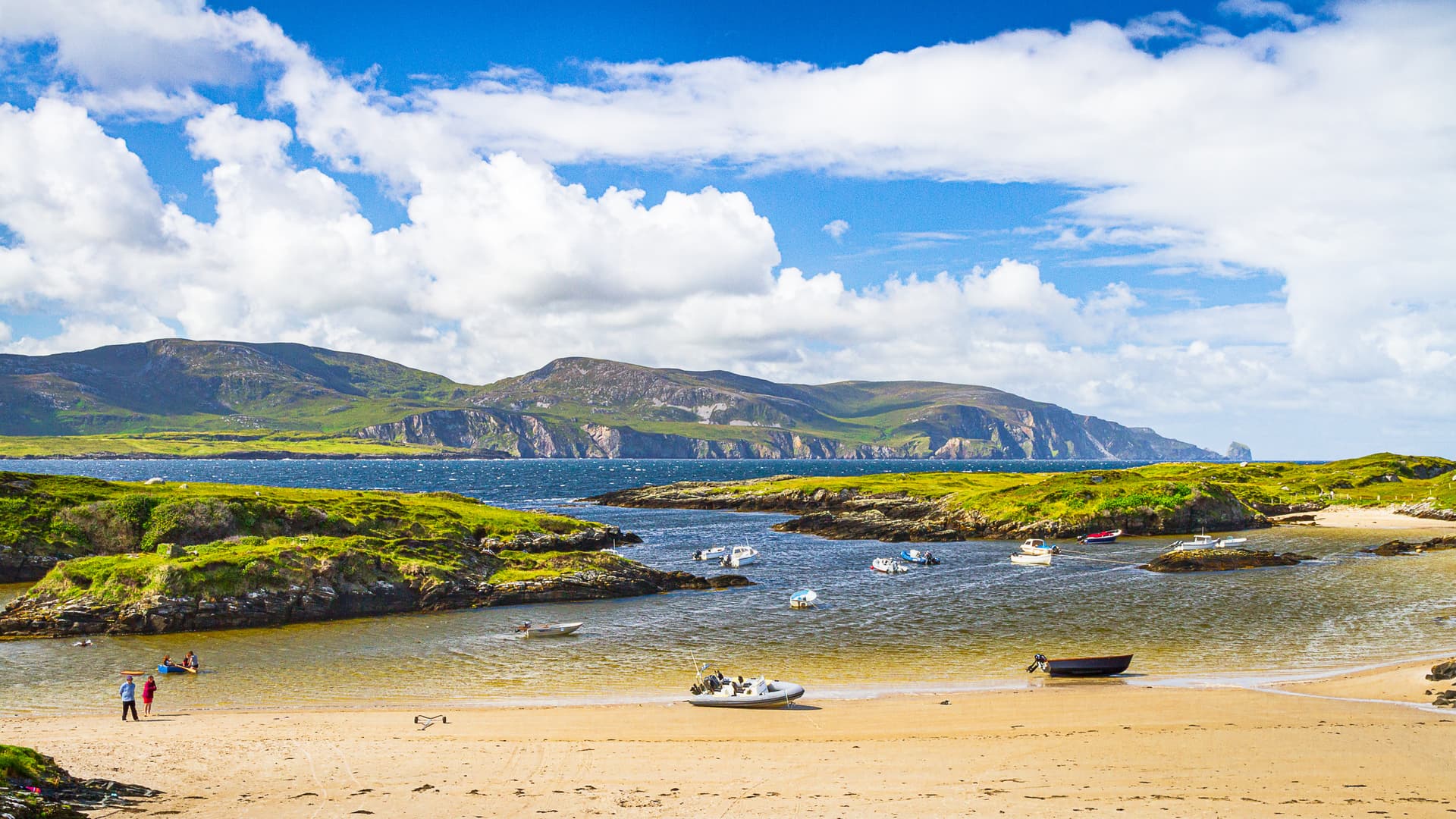 Small boats anchored in a cove with green hills and mountains under a blue cloudy sky.