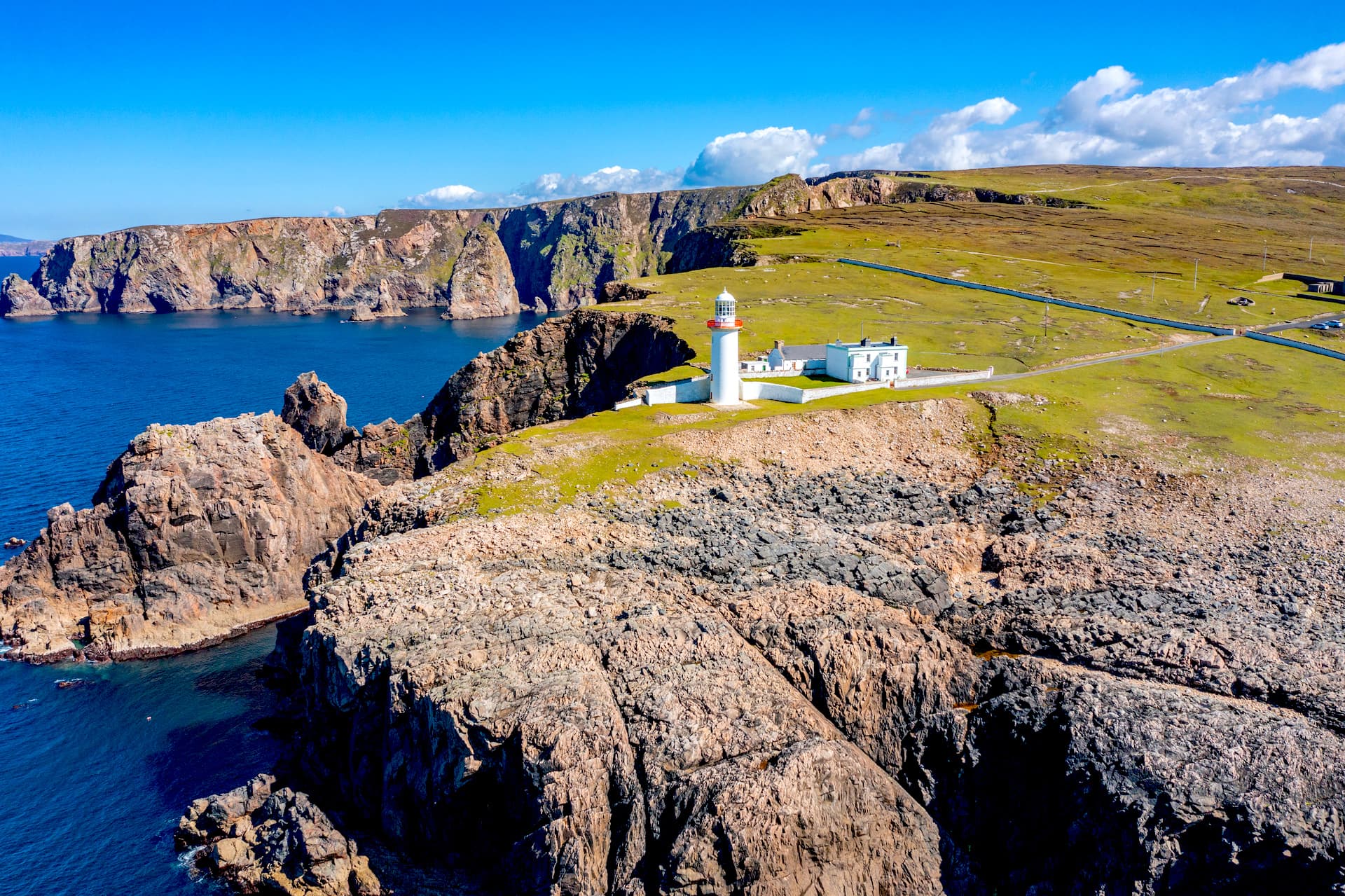 Arranmore Island lighthouse on grassy cliffs overlooking dark blue sea under bright blue sky.