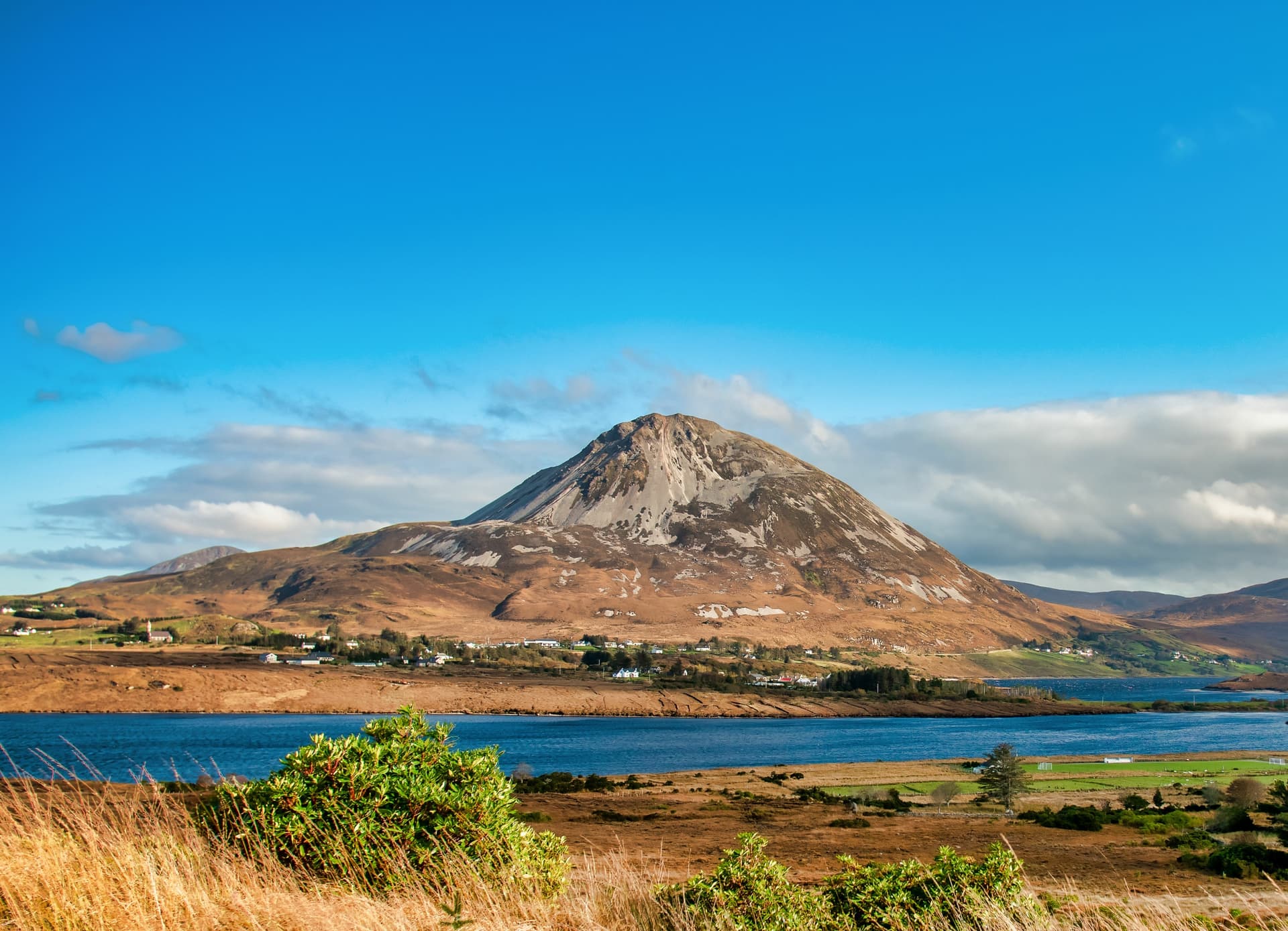 Mountain rising above blue water, village nestled in brown hills under bright blue sky.