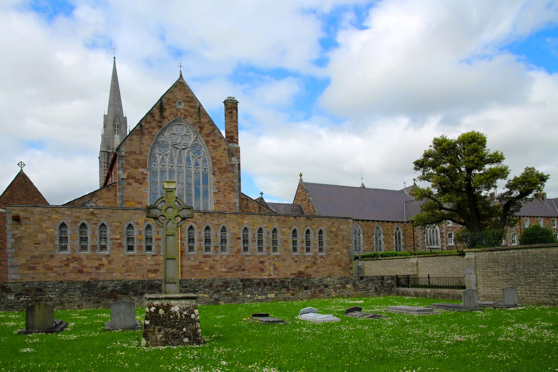 Stone Celtic cross in churchyard with Tralee church building and spire under cloudy sky.