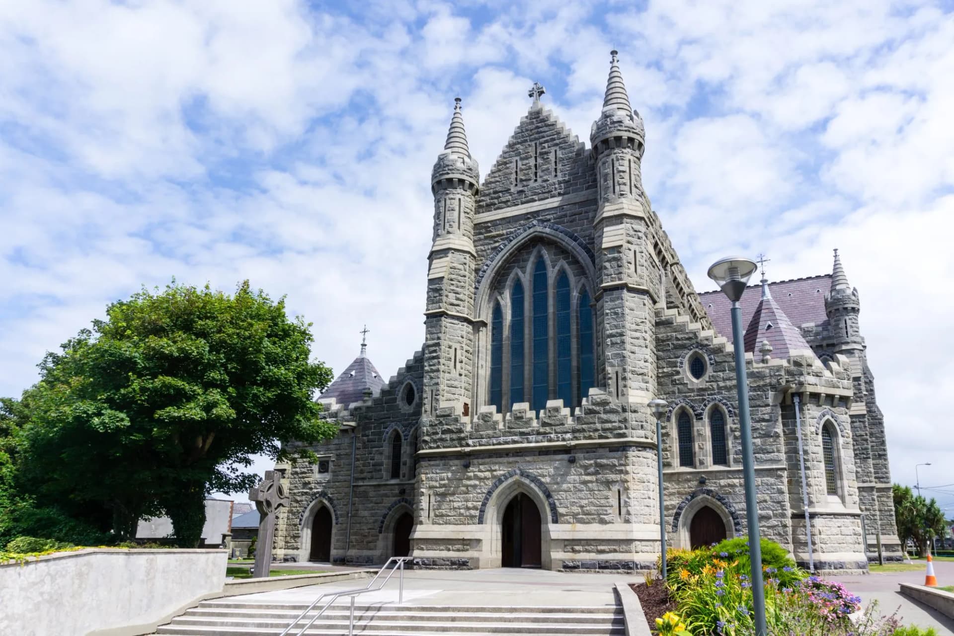 Stone church with turrets and large arched windows in Cahersiveen under a blue, cloudy sky.