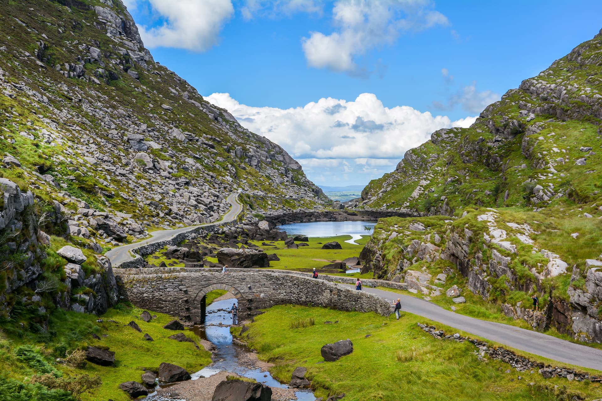 Gap of Dunloe stone bridge over stream, with winding road between green, rocky mountains.