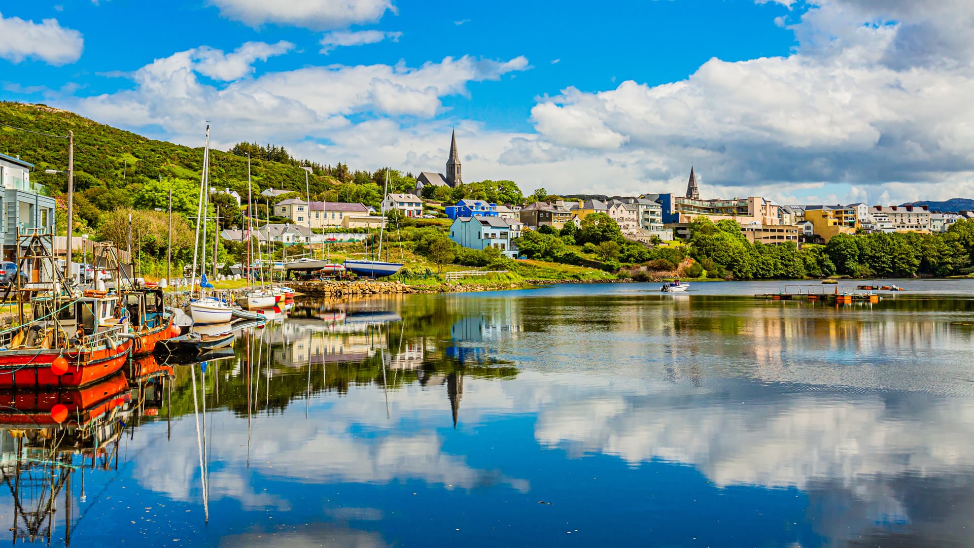 Boats docked in harbor reflecting on water near colorful town on green hillside in Clifden.