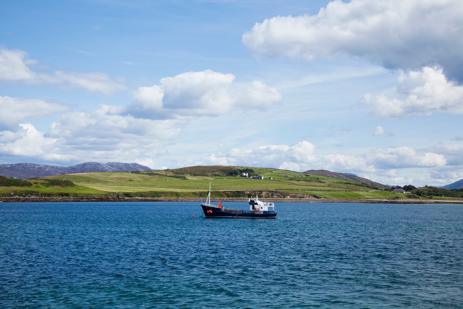 Small boat on blue water near green, rolling hills under a cloudy sky, Cleggan.