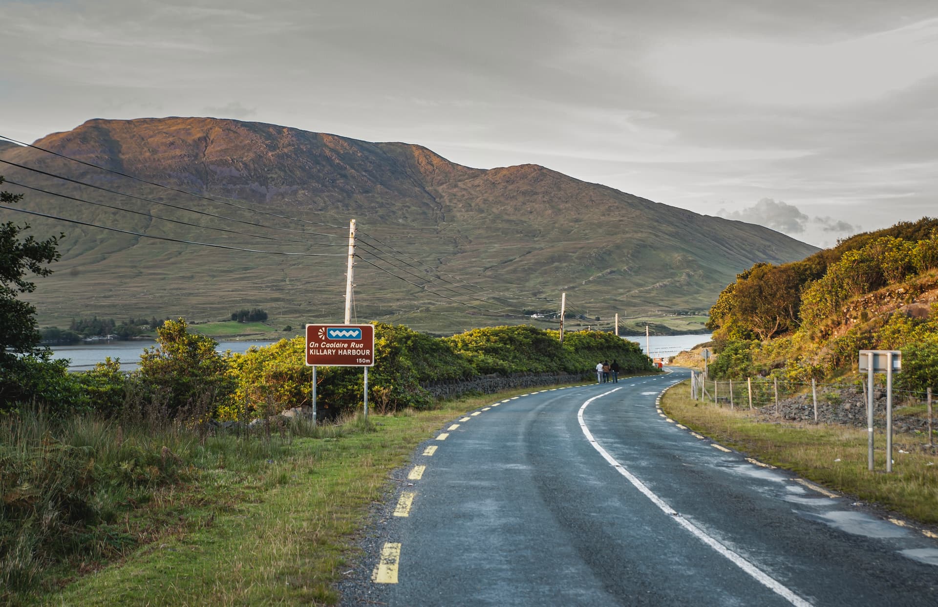 Winding road near Killary Harbour sign with large green mountain backdrop.