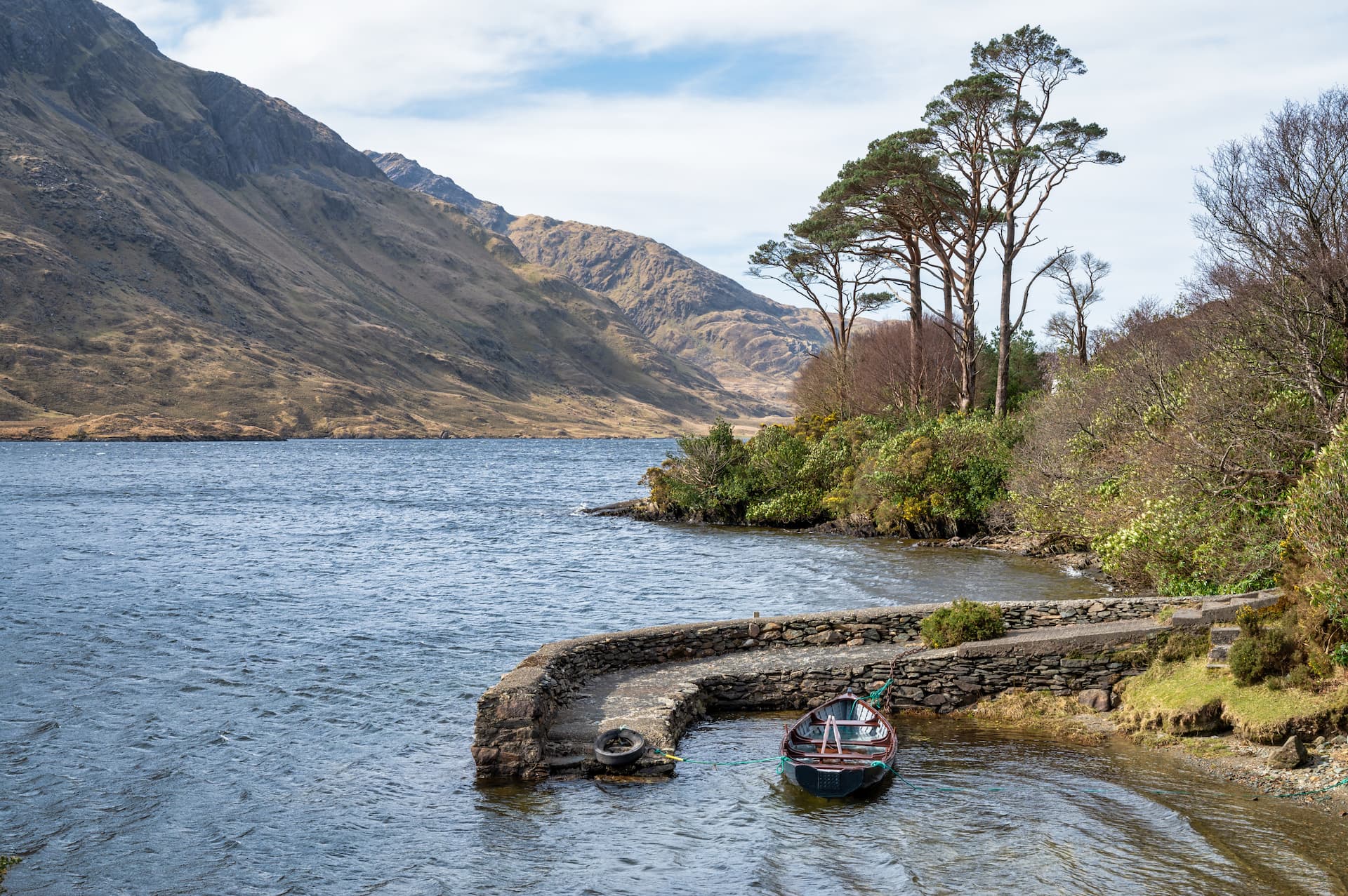 Small boat moored by stone quay on Doo Lough with steep, grassy mountains in background.