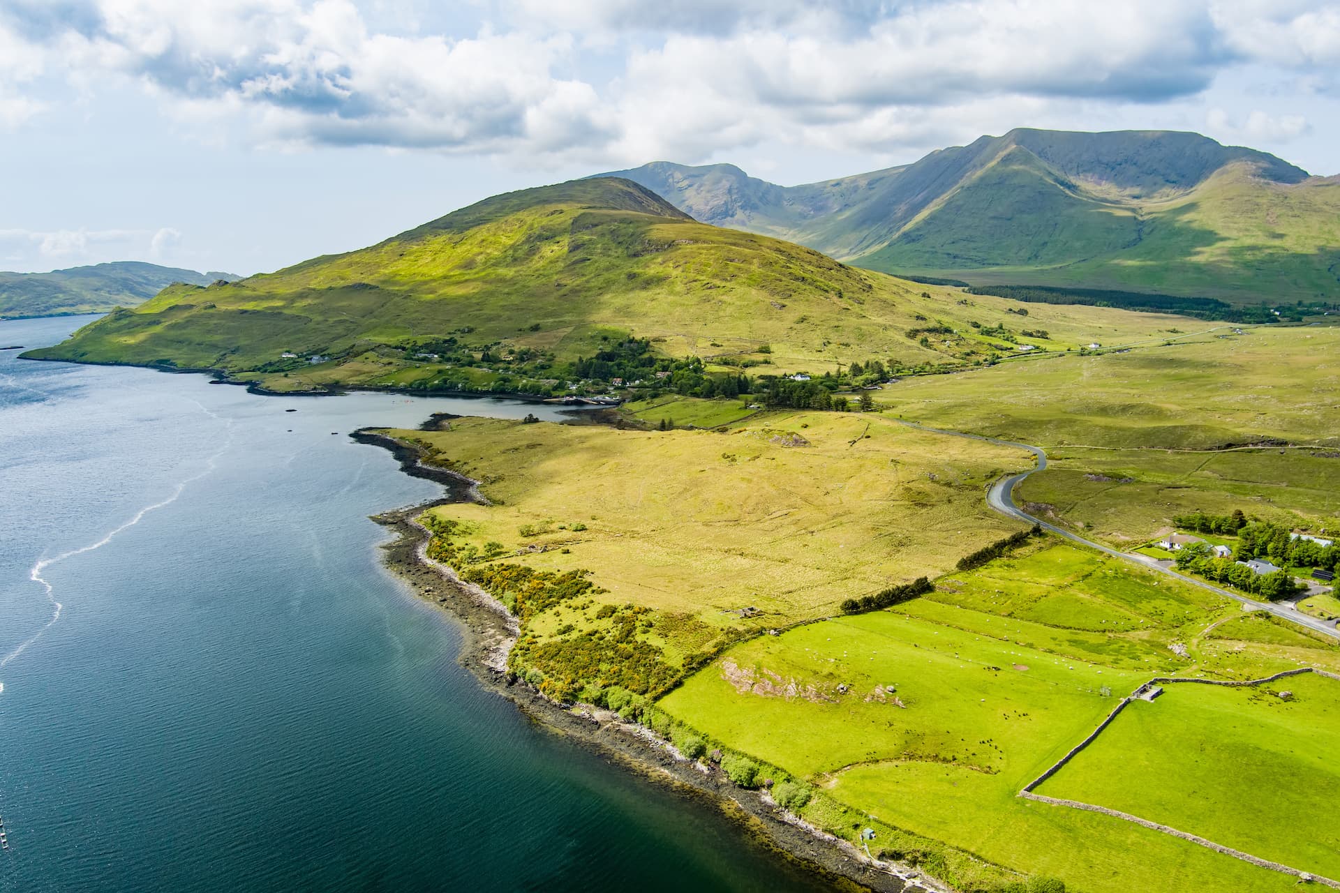 Aerial view of green rolling hills meeting dark blue coastal water under a cloudy sky at Killary Harbour.