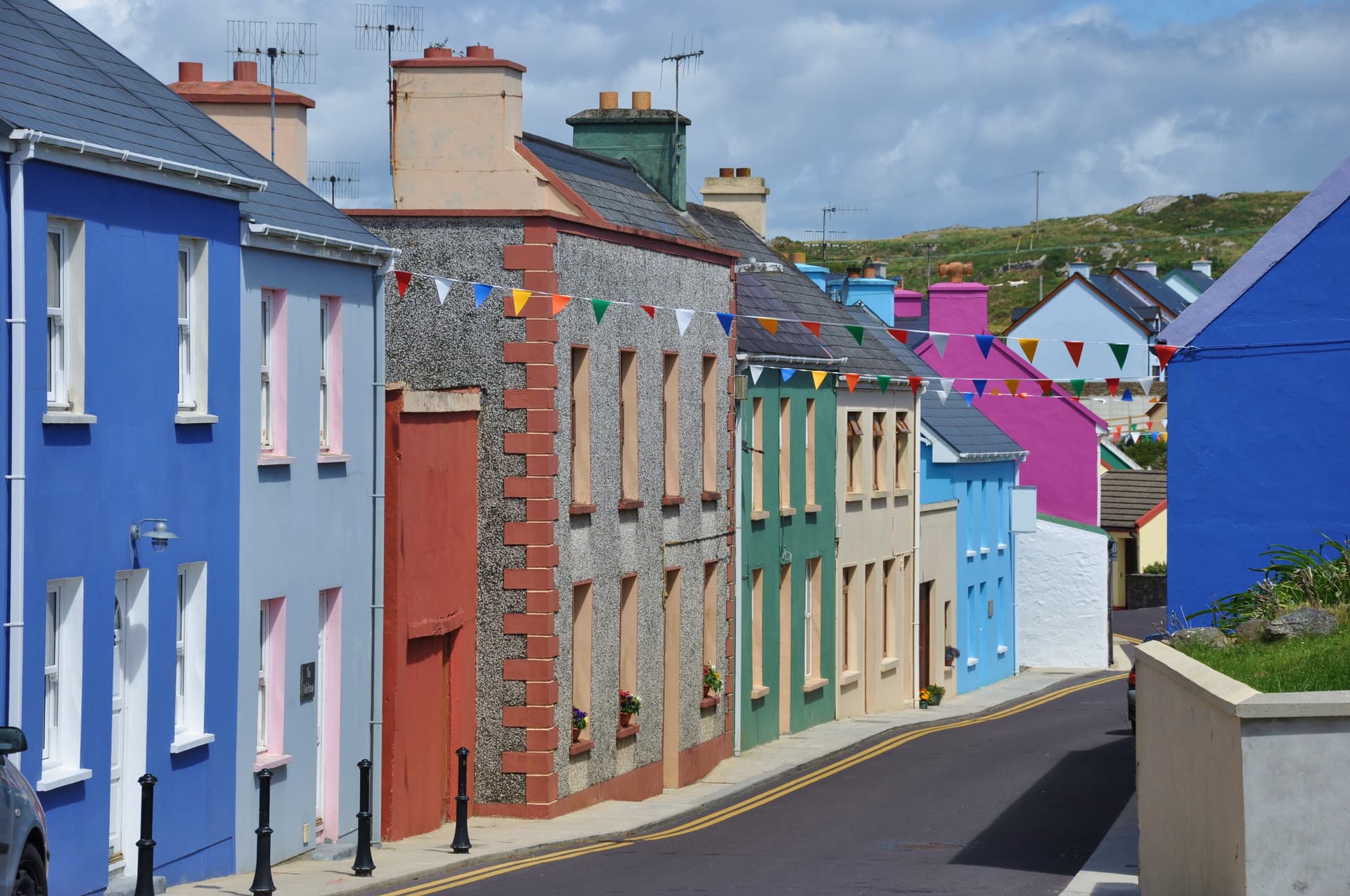 Colorful buildings line a street leading toward grassy hills under a cloudy sky.