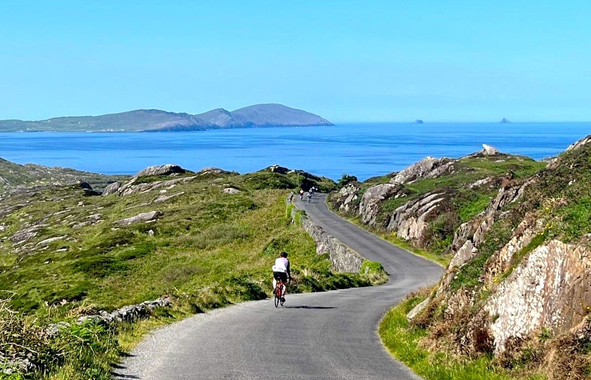 Cyclists riding on a winding coastal road with grassy hills and islands in the blue sea.