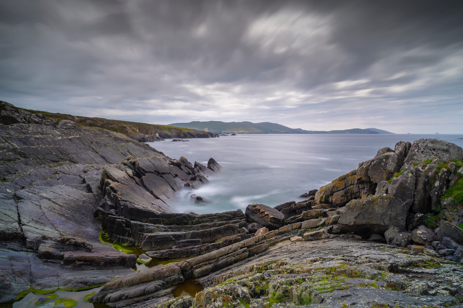 Rocky coastline with layered stone formations meeting the sea under a long exposure cloudy sky.