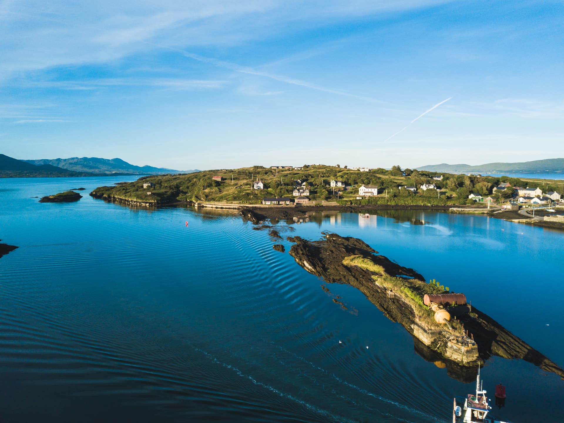 Coastal village on green hillside overlooking deep blue water with distant mountains under clear sky.