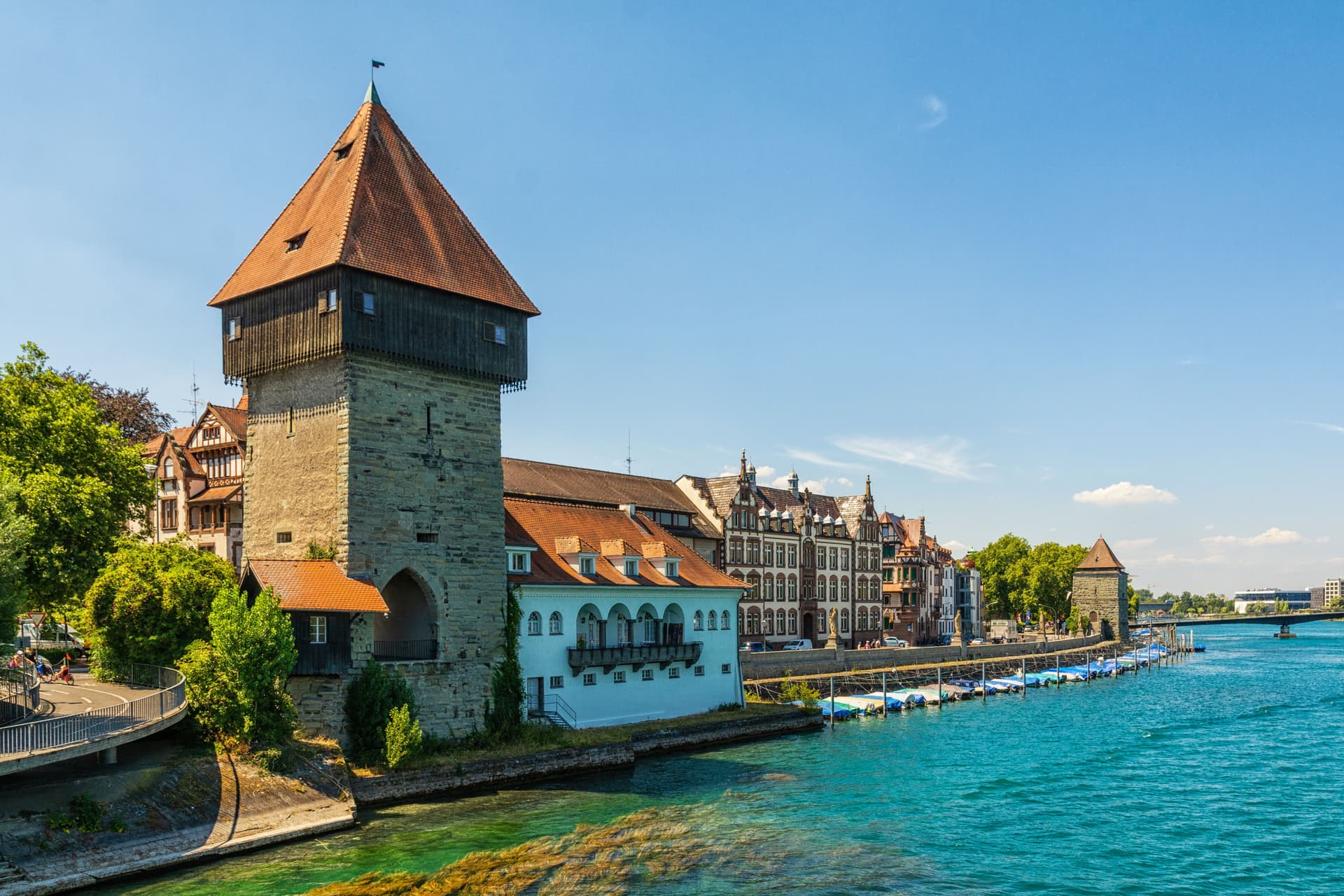 Stone tower with orange roof next to turquoise water and historic buildings in Constance.
