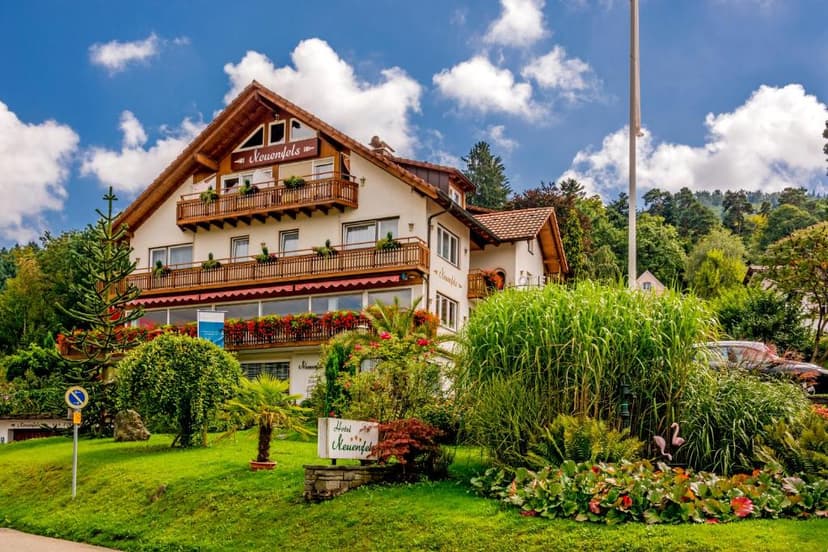 Hotel Neuenfels building with wooden balconies surrounded by lush green landscaping under a blue sky.