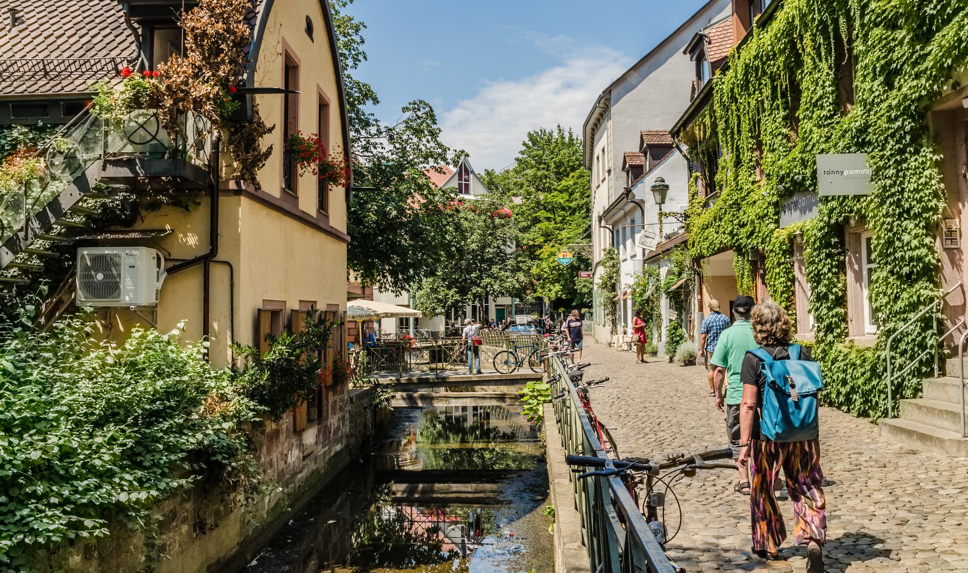 Pedestrians walk along a cobblestone street beside a small canal in Freiburg, Germany.