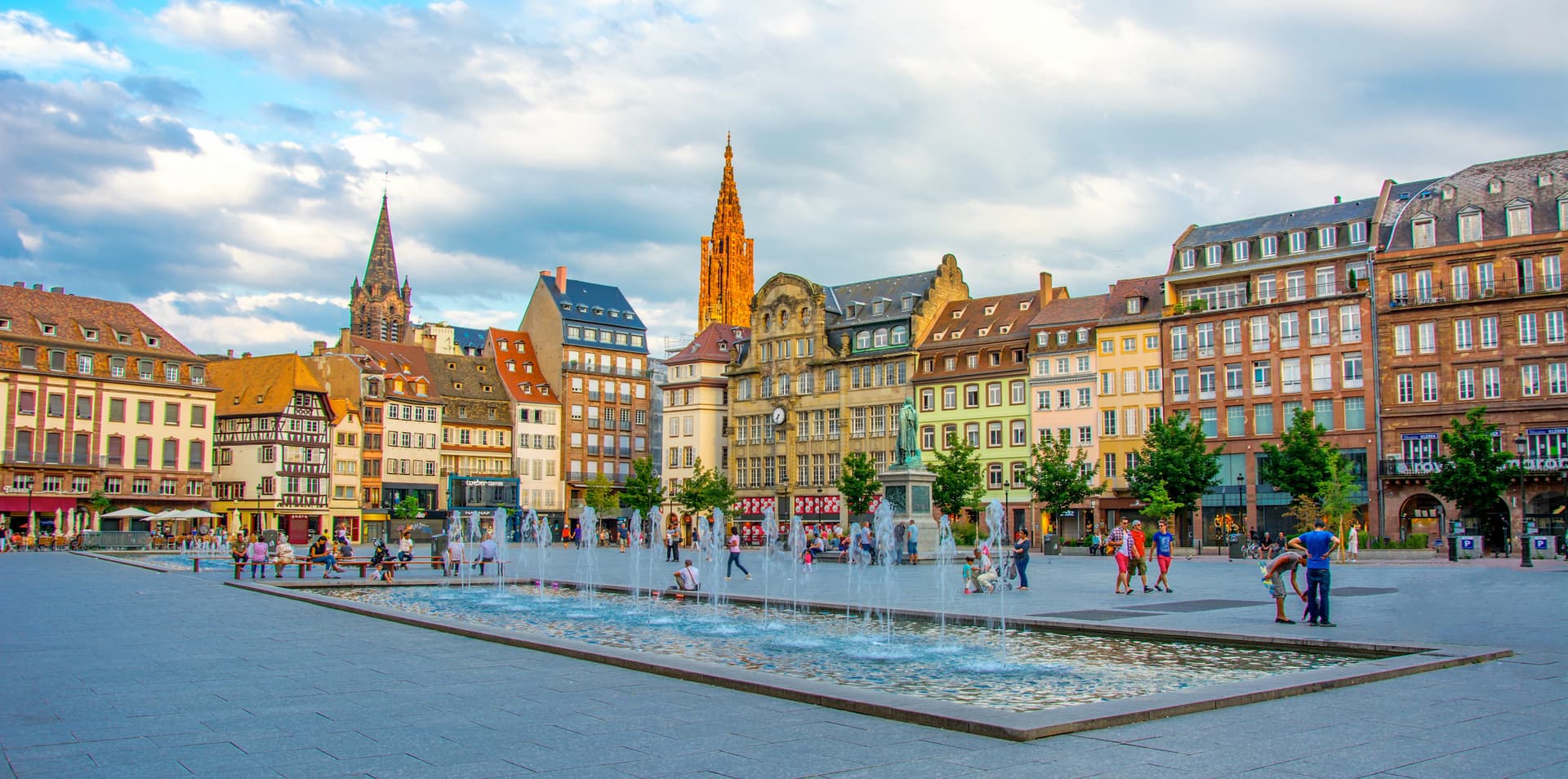 Fountain in Strasbourg square with historic buildings and church spire under cloudy sky.