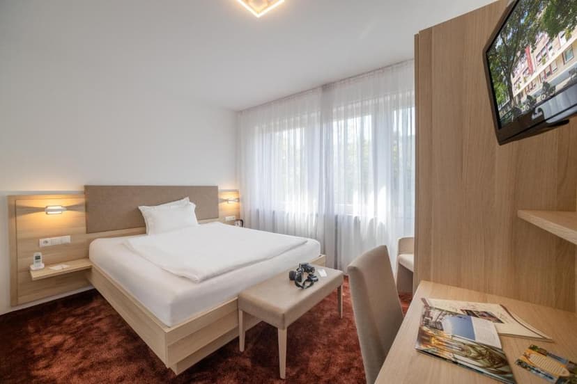 Modern hotel room with white bedding, wood accents, and sheer window curtains in Koblenz.
