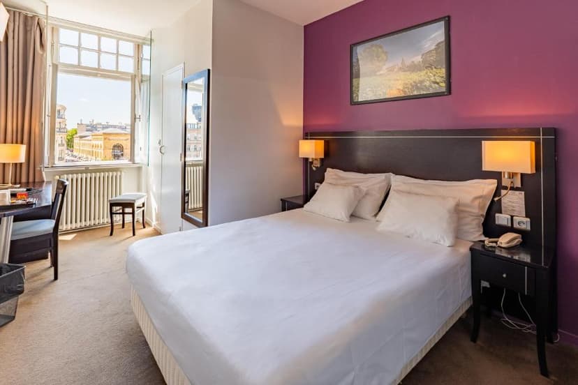 Hotel room with white bedding, purple accent wall, and view of European city buildings.