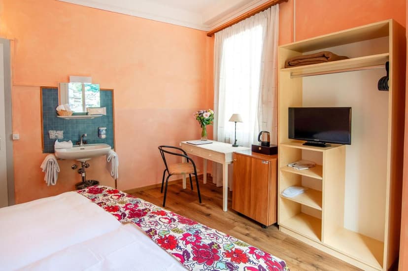 Simple hotel room with peach walls, open shelving, TV, desk, and sink area. Biasca.