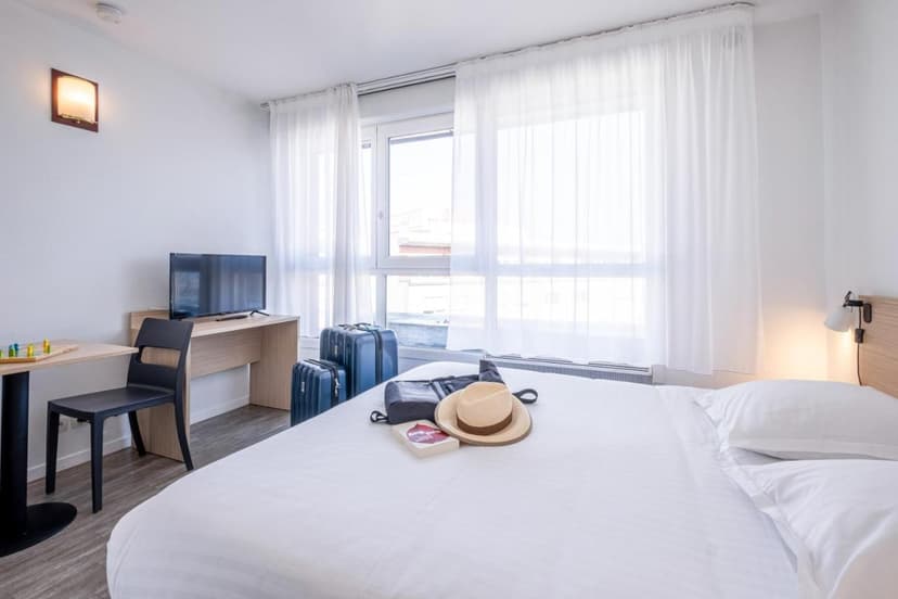 Bright hotel room with white bedding, luggage, TV, and travel hat near a large window.