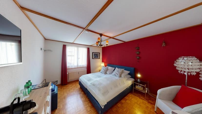 Bedroom with blue bed, red accent wall, wood floor, and white lamp at Buchs BnB Haus Weibel.