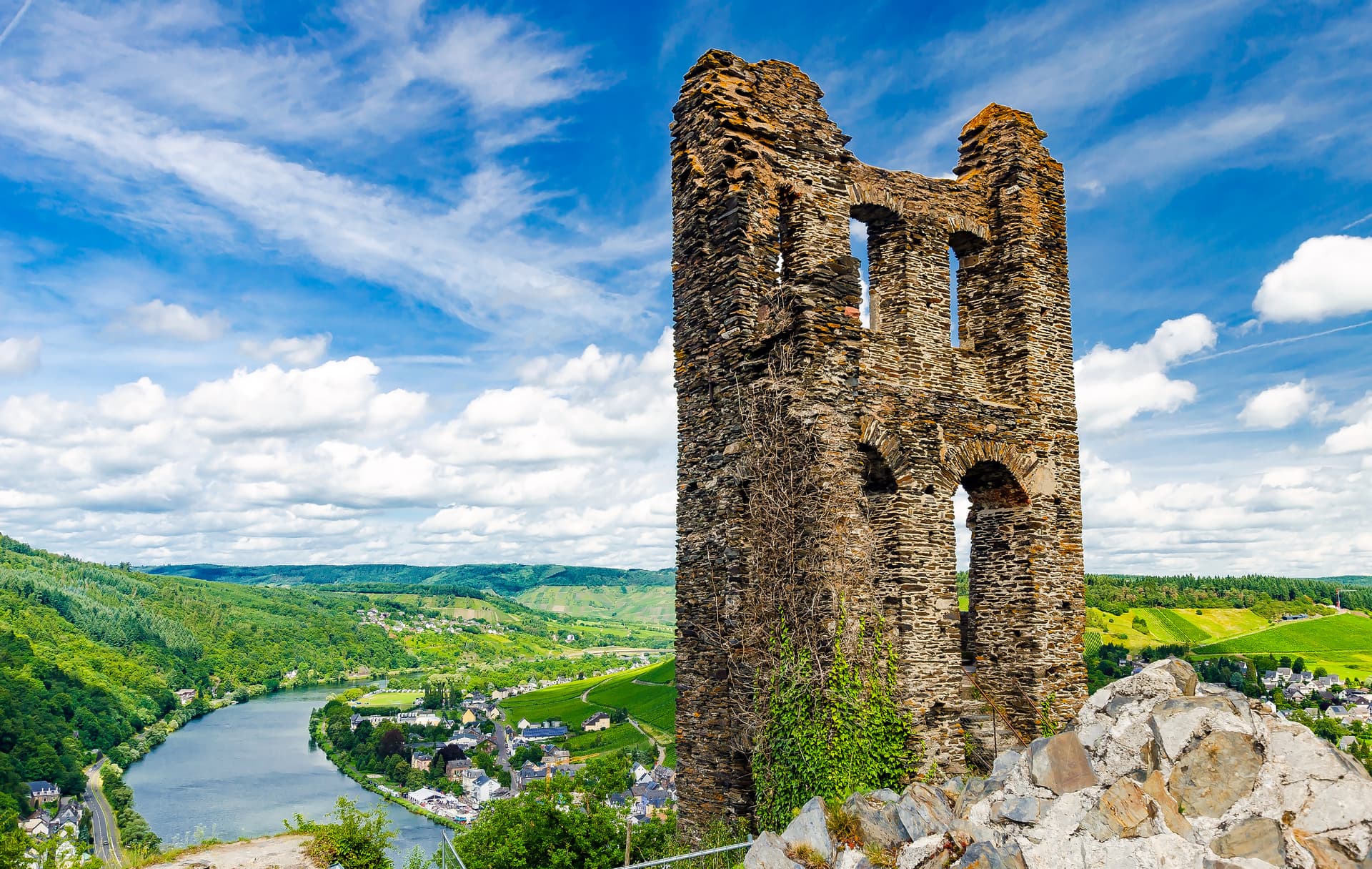 Stone castle ruins overlooking a town, river, and green vineyards under a blue sky.