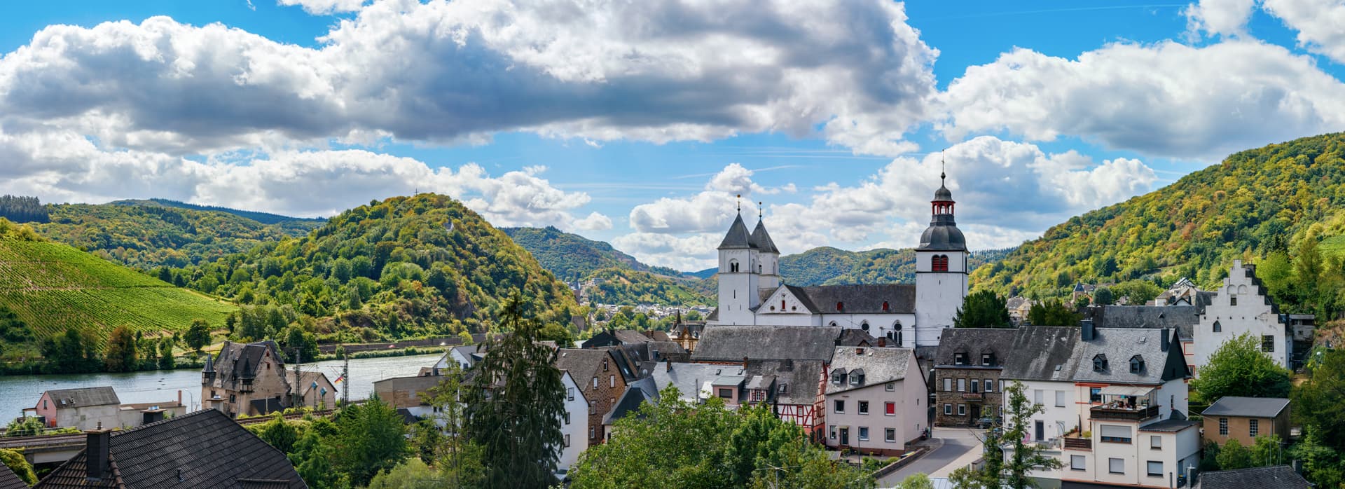 Town with white church towers nestled in green hills above a river under a cloudy blue sky.