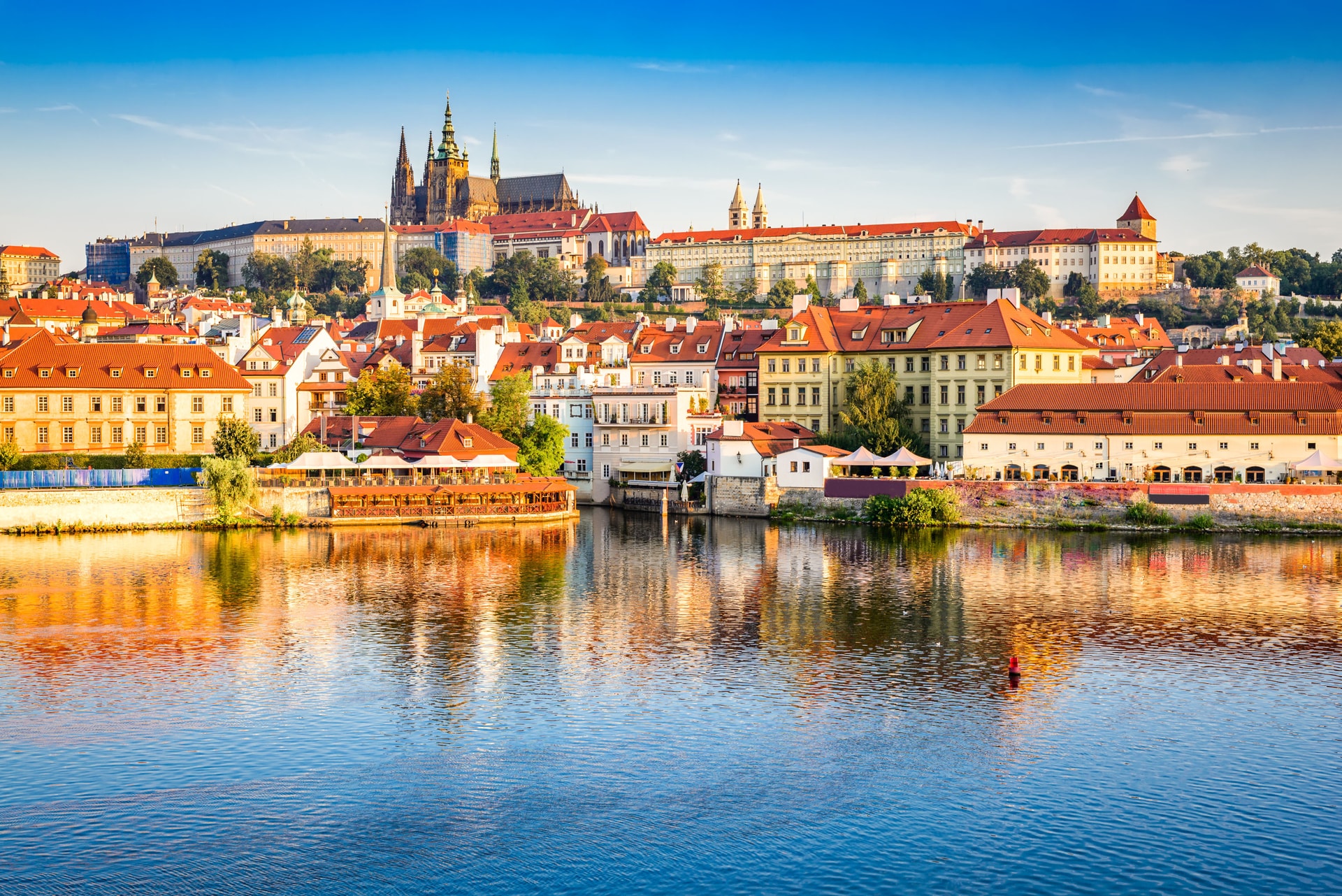 Prague Castle complex above colorful riverside buildings reflected in the Vltava River.