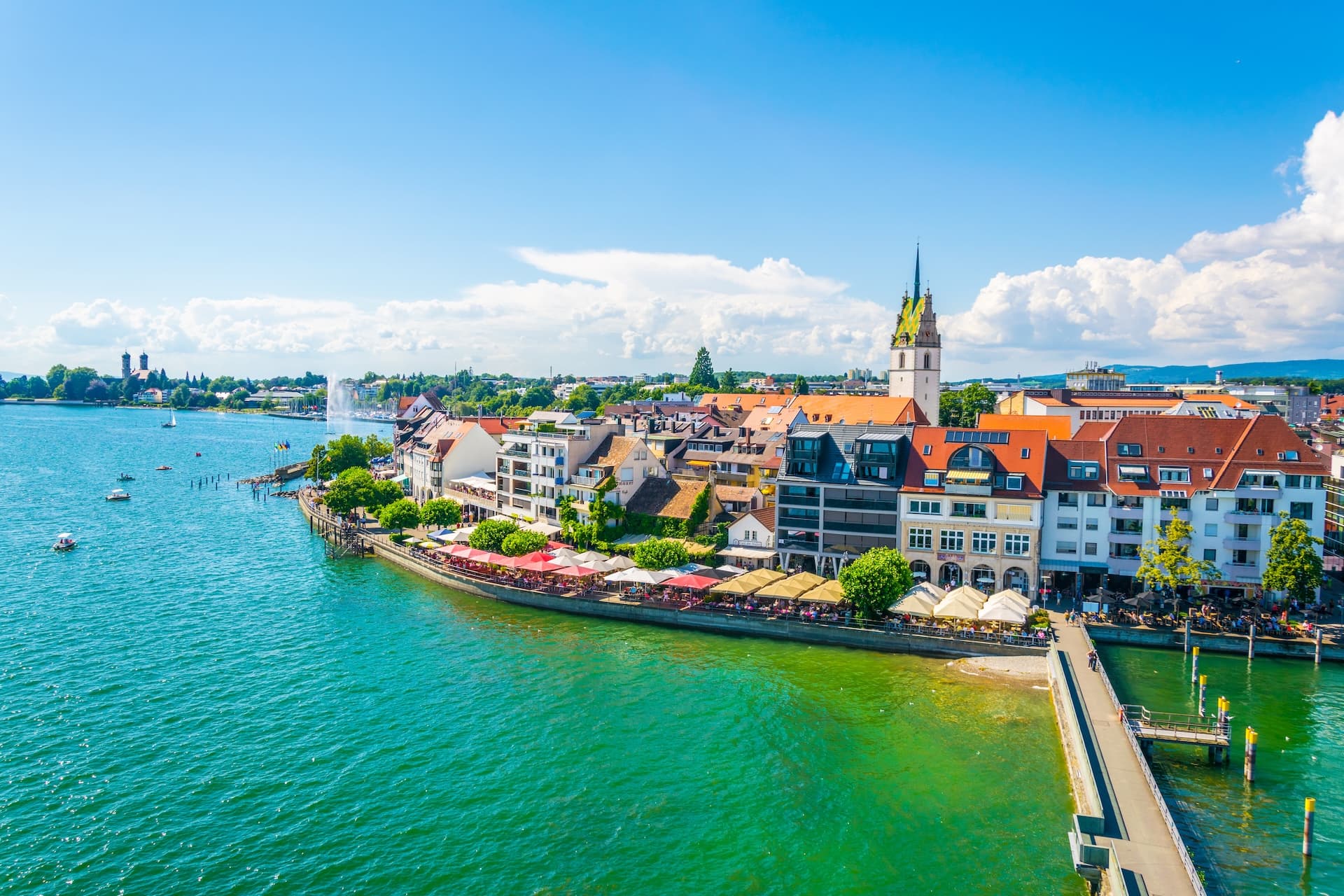 Panorama view of Friedrichshafen marina with colorful buildings and outdoor cafes along the green water.