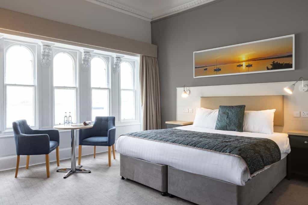 Hotel room with large bay windows, double bed, and seating area; sunset boat art above bed.