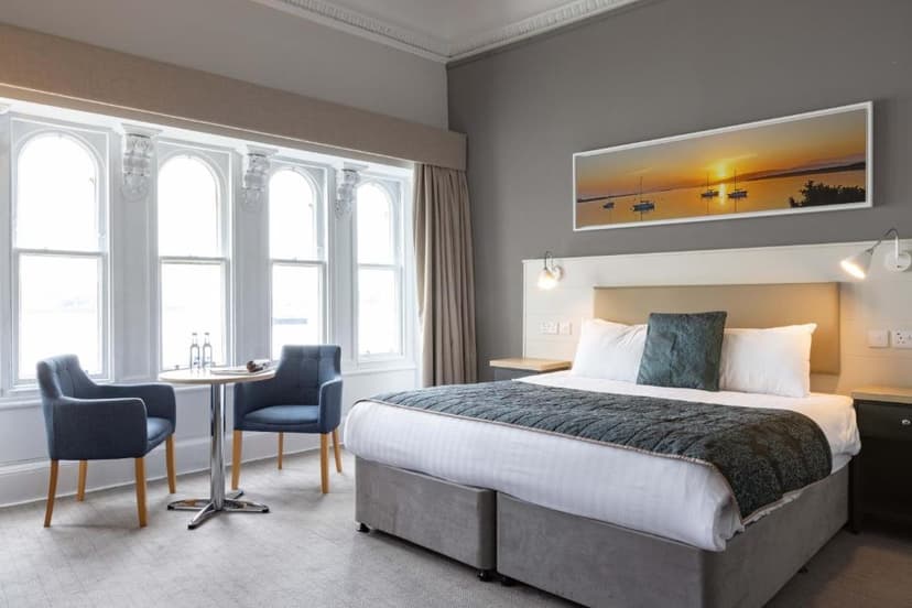 Hotel room with large bay windows, double bed, and seating area; sunset boat art above bed.