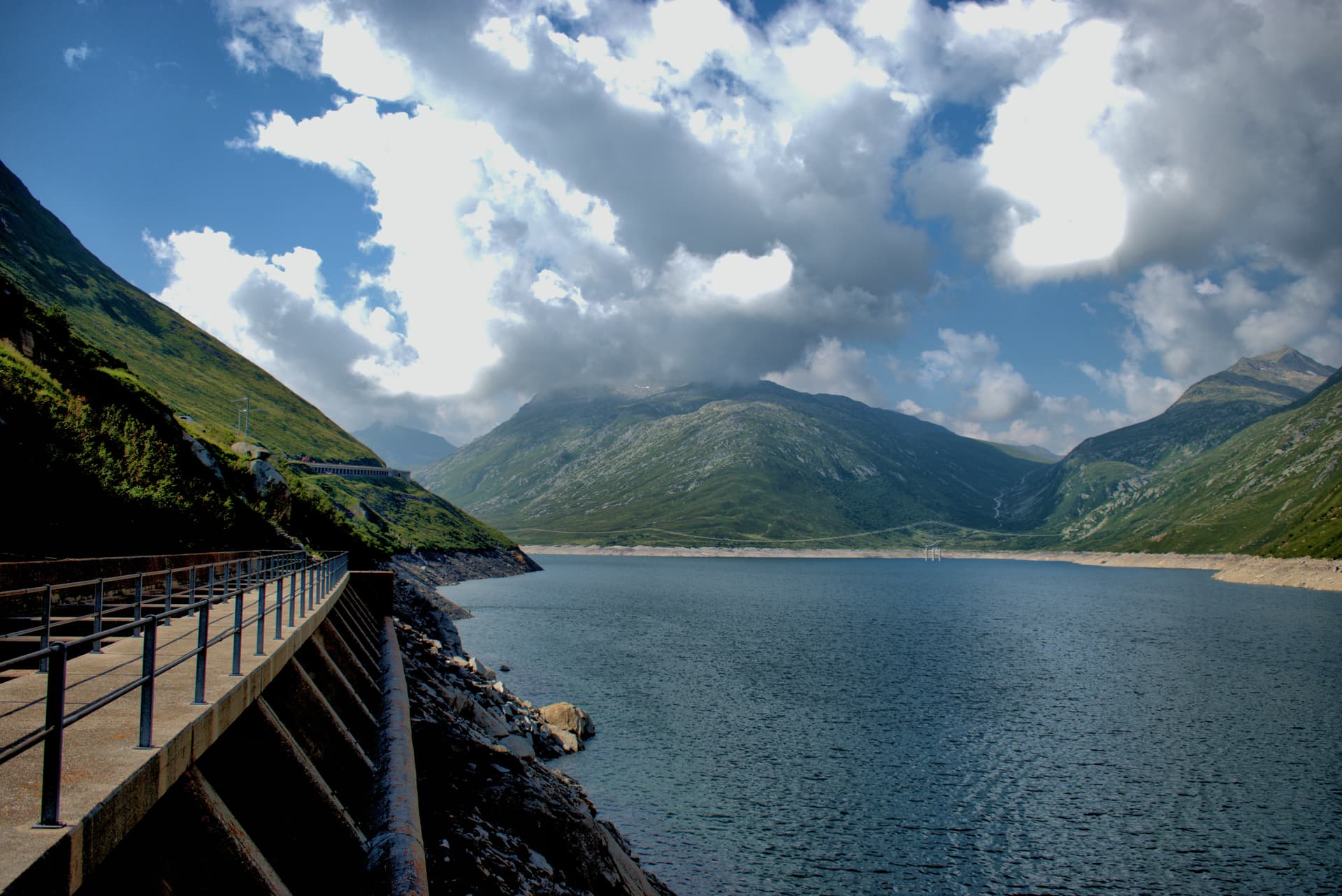 Dam walkway overlooking a reservoir surrounded by green alpine mountains under a cloudy blue sky at Lukmanierpass.