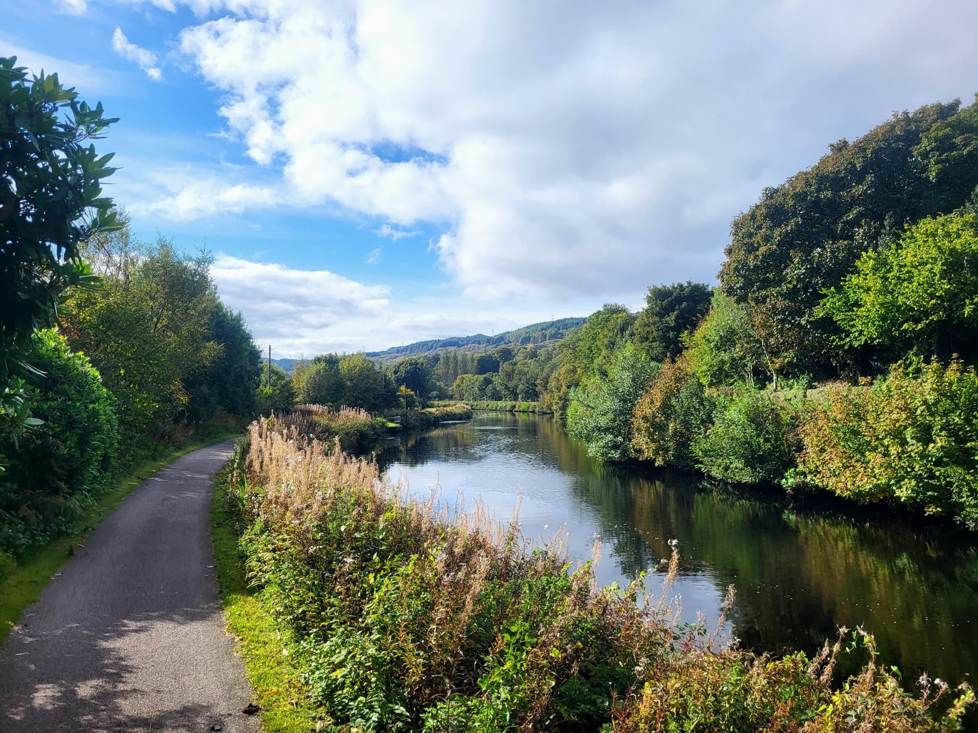 Crinan Canal towpath alongside dark water reflecting trees under a partly cloudy blue sky.