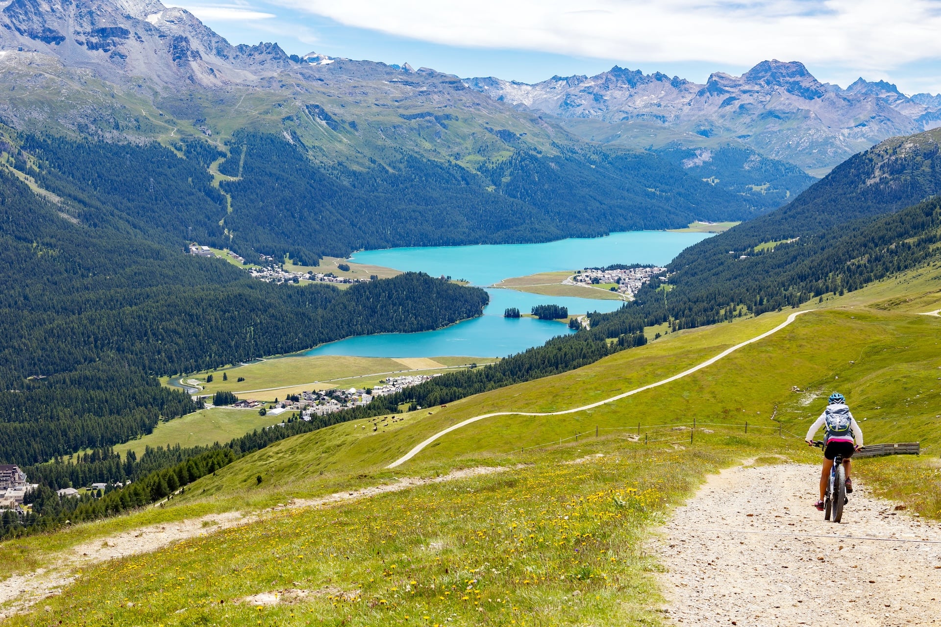 Mountain biker descending dirt trail overlooking turquoise lakes and alpine village in Sankt Moritz, Switzerland.
