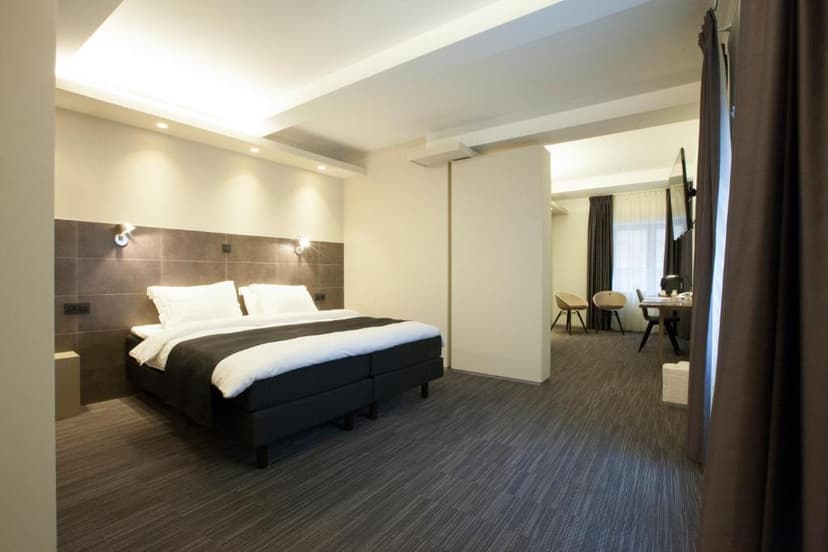 Modern hotel room with large bed, dark carpet, and separate seating area visible through doorway.