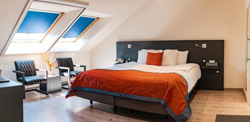Attic bedroom with sloped ceiling, skylights, large bed with orange duvet, and two dark armchairs.