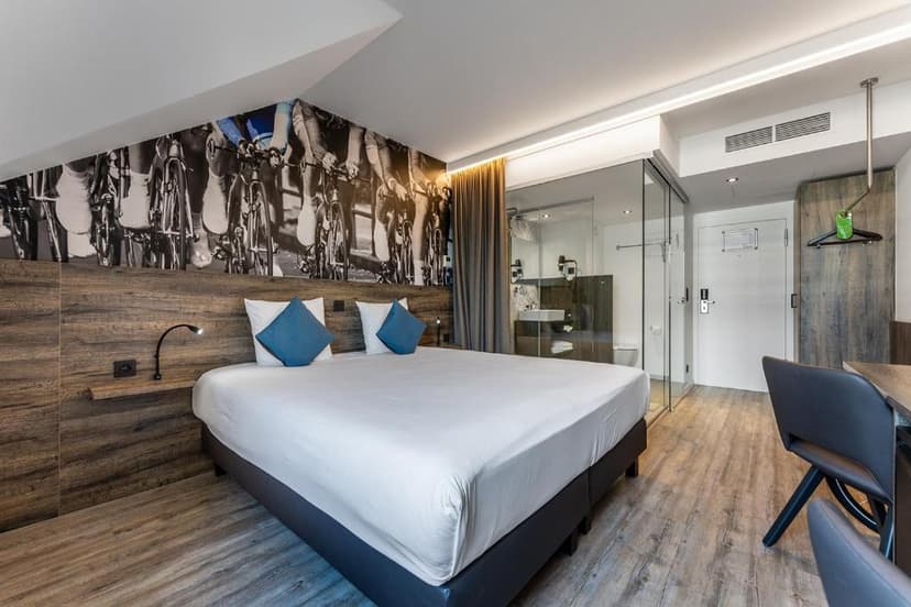 Modern hotel room with large bed, cycling mural, and glass-walled bathroom.
