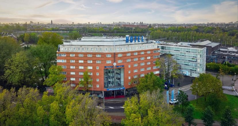 Hotel building with "HOTEL" sign surrounded by green trees, overlooking a distant city skyline.