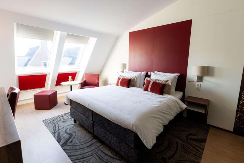 Hotel attic bedroom with white bedding, red accents, and sloped windows showing rooftops.