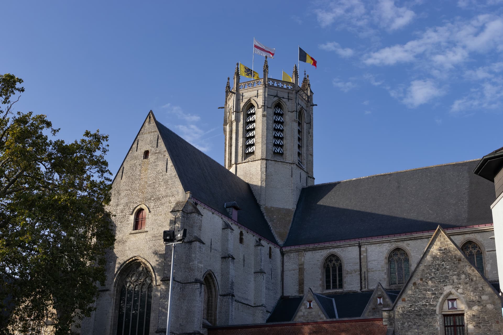 Stone church tower with flags flying under a clear blue sky in Dendermonde.
