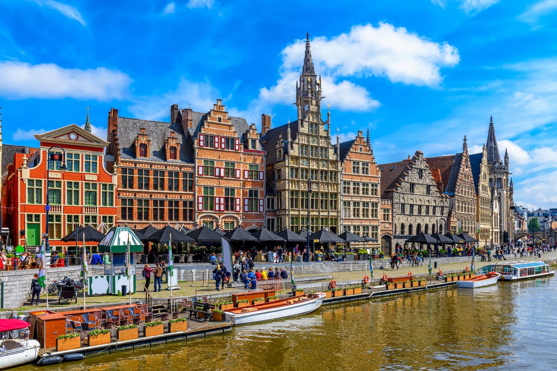 Historic guild houses line the canal in Ghent under a bright blue sky with white clouds.