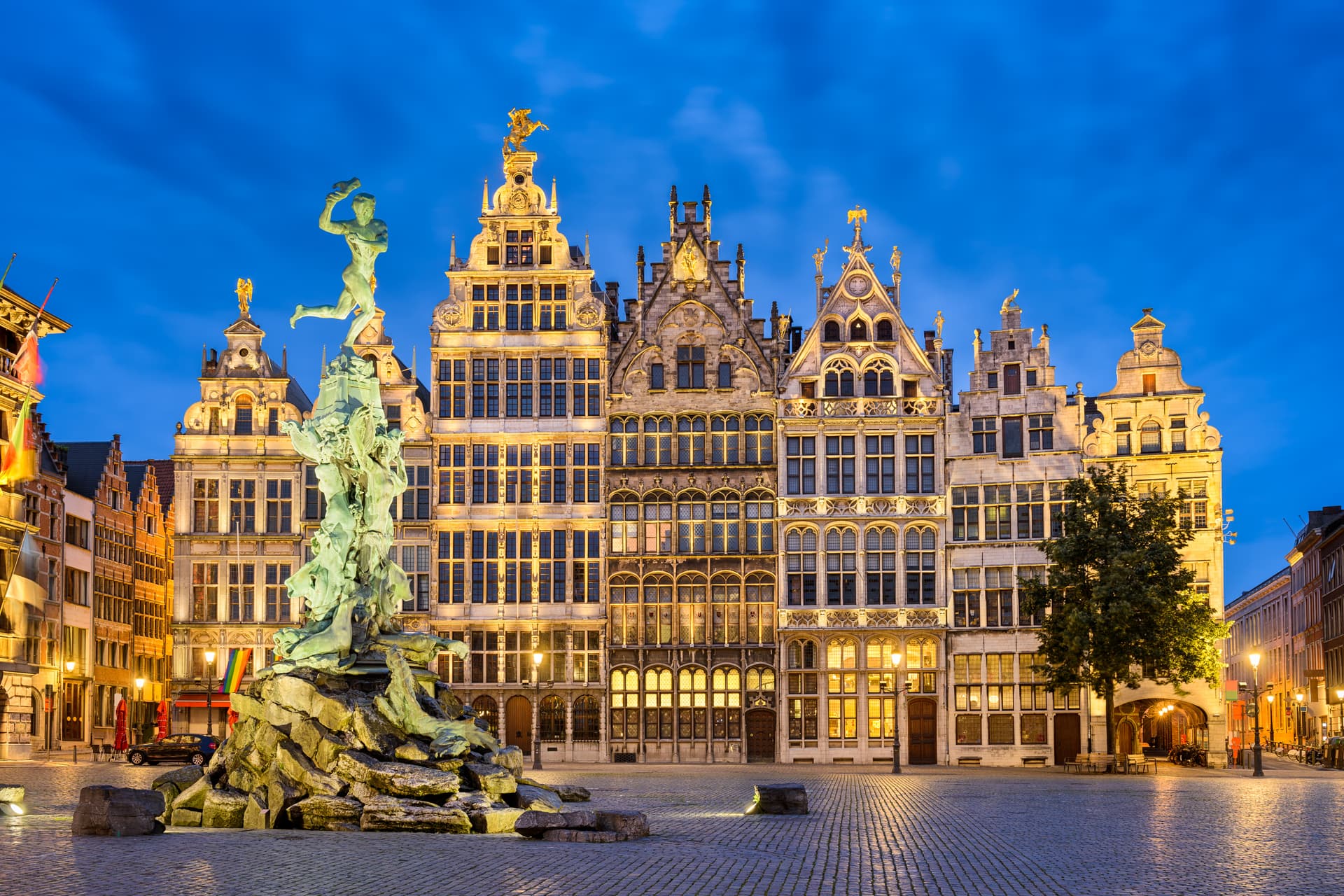 Statue of Brabo fountain in cobblestone square before illuminated guildhall buildings at twilight.